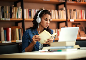Studentin lernt in einer Bibliothek mit Kopfhörern und Buch in der Hand am Laptop