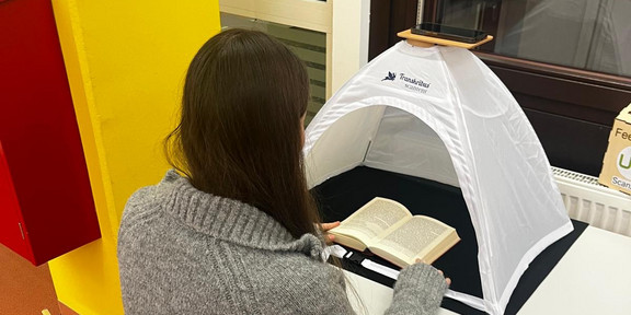 Junge Frau scannt mit ihrem Smartphone Seiten aus einem Buch ein, das unter einem Scanzelt liegt.