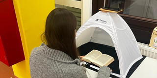 Junge Frau scannt mit ihrem Smartphone Seiten aus einem Buch ein, das unter einem Scanzelt liegt.