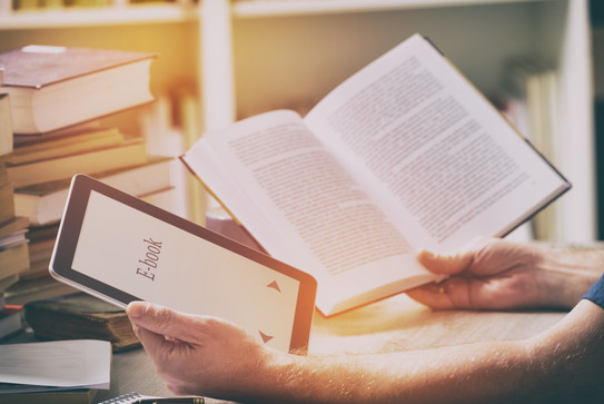 A man sits with an e-book reader and a book in his hands at a table with books, stationery and papers