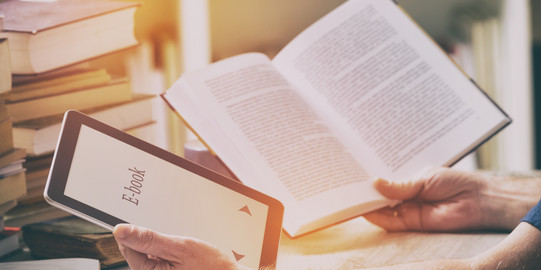 Ein Mann sitzt mit einem E-Book-Reader und einem Buch in den Händen an einem Tisch mit Büchern, Schreibzeug und Papieren