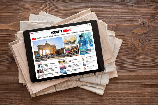 A tablet with a news website lies on a stack of newspapers