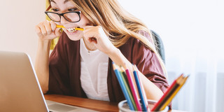 A young woman sits in front of a laptop while biting a pencil