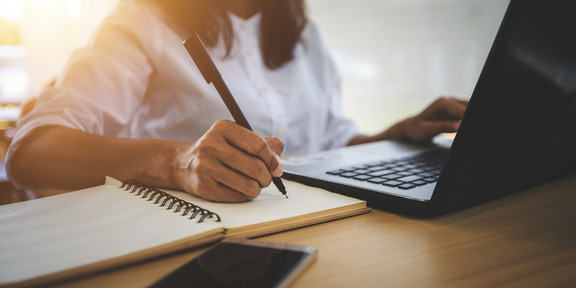 A young woman learns on a laptop and writes in a notebook