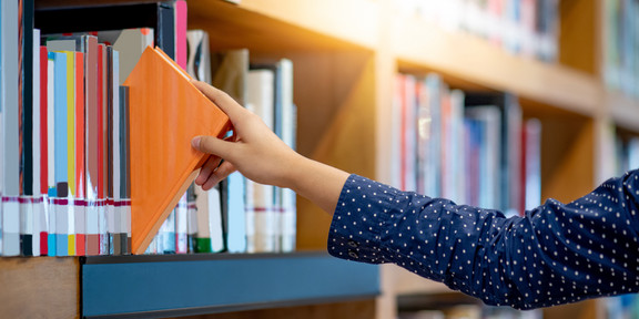 A right hand pulls out an orange book from a wooden shelf. 