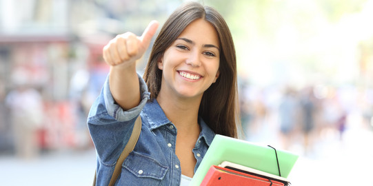 Eine fröhliche Studentin mit Heften im Arm zeigt "Daumen hoch"