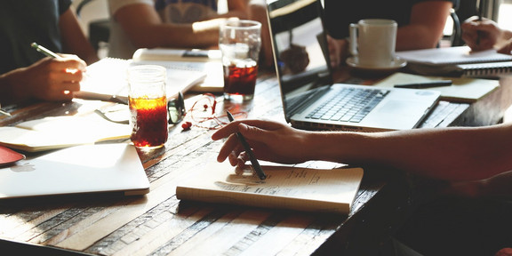 Schreibende Personen sitzen an einem Tisch mit Laptop, Heften, Getränken