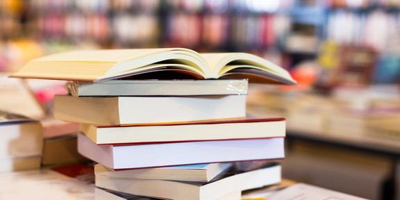 Unordentlicher Bücherstapel liegt auf einem Tisch, im Hintergrund Buchregale