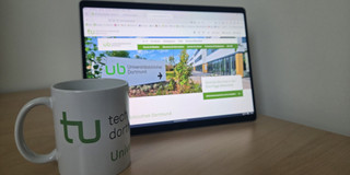Eine Tasse mit TU-Logo steht vor einem Bildschirm.