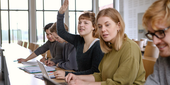 Studierende Laptops und Handmeldung Studierende im Seminarraum sitzen in einer Tischreihe vor ihren Laptops. Eine Studierende meldet sich.