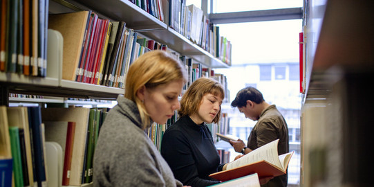 Book research in the library Students stand reading in front of bookshelves in the library.