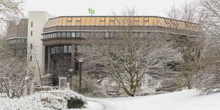 Bare trees, snow-covered green spaces, the library building in the background and the TU logo in winter.