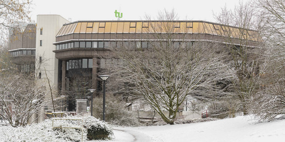 Gebäude der Bibliothek im Schnee Kahle Bäume, verschneite Grünflächen, im Hintergrund das Bibliotheksgebäude und das TU-Logo im Winter.