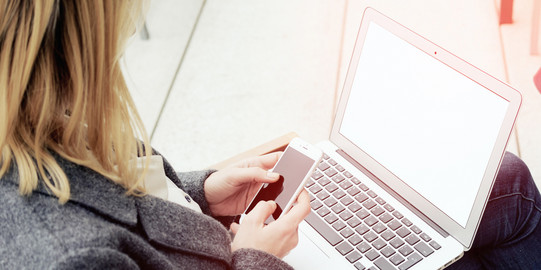 Frau mit Laptop auf dem Schoß, hält ein Smartphone in den Händen.