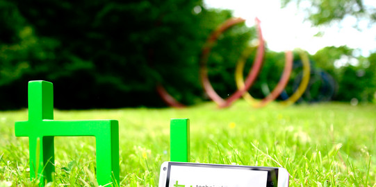 Ein Smartphone, das neben dem TU-Logo im Gras steht, im Hintergrund sind die Spektralringe zu sehen.