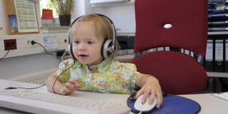 A toddler wearing headphones is sitting in front of a computer.