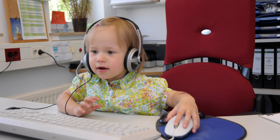 Children_03 A toddler wearing headphones is sitting in front of a computer.