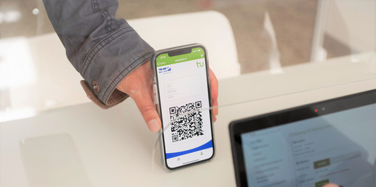 One hand holds a smartphone with a QR code in the TU app, which is scanned by a tablet.
