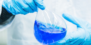 Two hands in blue rubber gloves hold a volumetric flask filled with blue liquid.