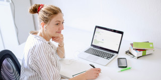 Woman sitting at laptop Woman sitting at her desk with a laptop writing something down in a notebook